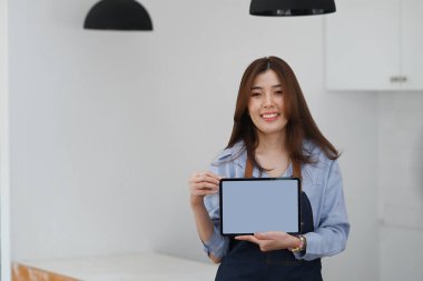 Önlüklü başarılı küçük işletme sahibi boş ekranlı dijital tableti gösteriyor ve kameraya gülümsüyor.