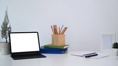 Bilgisayar tableti, bitki, defter, kalem tutacağı ve beyaz masa üzerine kitabı olan şık bir iş yeri.