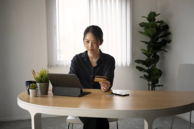 Genç Asyalı kadın dijital tablet kullanıyor ve çevrimiçi ödeme veya ofisteki online alışveriş için kredi kartı tutuyor.