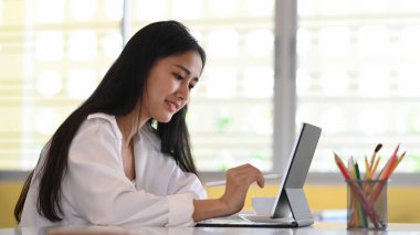 Çekici genç tasarımcı rahat bir iş yerinde oturup bilgisayar tableti üzerinde çalışıyor..