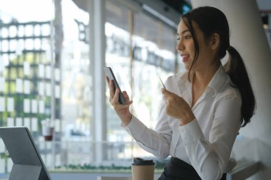 Gülümseyen Asyalı genç kadın cep telefonu kullanıyor ve çevrimiçi ödeme veya online alışveriş için kredi kartı tutuyor.