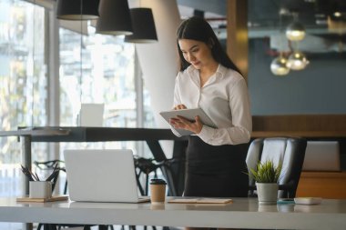 Çekici kadın ofis çalışanı dijital tablet tutuyor ve iş yerinde duruyor..
