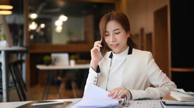 Kendine güveni tam bir iş kadını. Belgeleri kontrol ediyor ve asistanına cep telefonundan talimatlar veriyor..