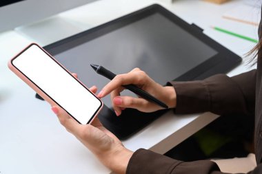 Dijitalleştirici kalem tutan ve iş yerinde cep telefonu kullanan kadın grafik tasarımcısının görüntüsünü kapat.