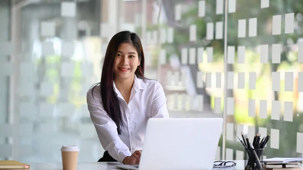幸せな若い女性の従業員は ラップトップコンピュータと現代のオフィスに座って カメラに笑顔 — ストック写真
