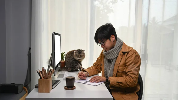 Young man sitting in front of computer with his cat and making note on notebook at home office.