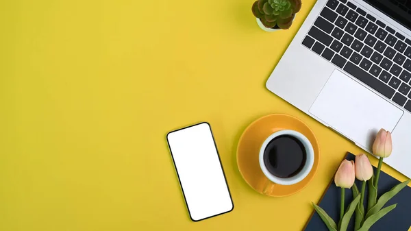 Top view of female workspace with computer laptop, coffee cup, flowers, smart phone and copy space on yellow background.