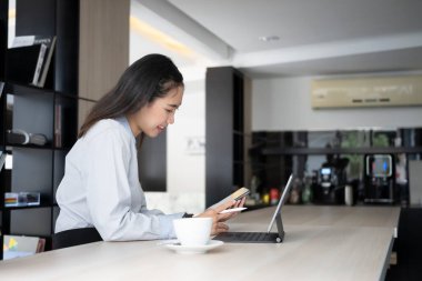 Ofisteki bilgisayar tabletinin önünde oturan ve akıllı telefon kullanan genç bayanın yan görüntüsü.