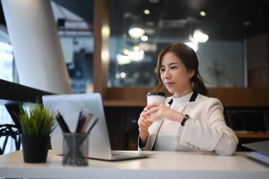 Başarılı bir iş kadını elinde kahve fincanı tutuyor ve modern iş yerindeki dizüstü bilgisayardan bilgi okuyor..