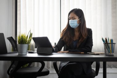 Koruyucu maskeli bir iş kadını ofis masasında bilgisayar tableti üzerinde çalışıyor..