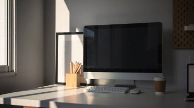 Pencerenin yanındaki beyaz masada bilgisayar ve ofis malzemeleri olan modern ev ofisi..