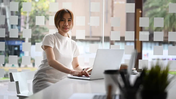 自信を持っている女性起業家は 彼女のラップトップコンピュータと現代のワークスペースに座って カメラに笑顔 — ストック写真