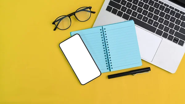 Top view of creative workspace with, computer laptop, notebook, smart phone and glasses on yellow background.