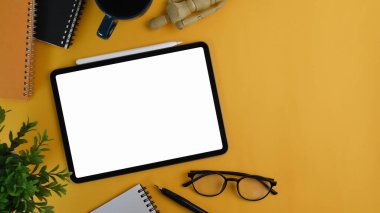 Mock up digital tablet with white screen, coffee cup, house plant ,glasses and notebook yellow background.