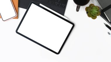 Top view of digital tablet with empty screen, coffee cup, glasses, notebook and house plant on white office desk.