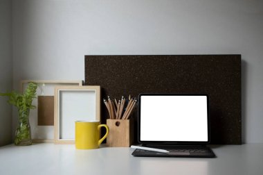 Kablosuz klavye, kahve fincanı, kalem tutacağı, ev bitkisi ve resim çerçeveli dijital tablet beyaz masa üzerinde.