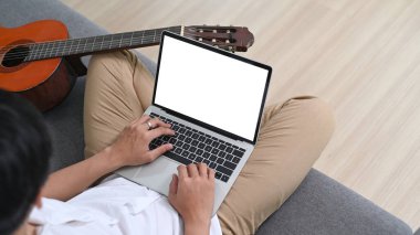 Casual man using laptop computer while sitting with his guitar on sofa at home.