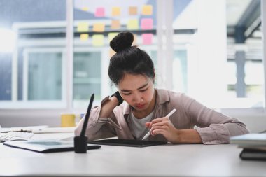 Genç bayan tasarımcı yaratıcı ofiste otururken dijital tablet üzerinde çalışmaya konsantre oluyor..