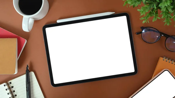 Digital tablet with white screen, glasses, coffee cup, notebook and houseplant on brown leather.