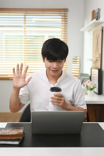 幸せな若いアジアの男を保持コーヒーカップと挨拶彼の友人上のビデオ通話上のラップトップコンピュータ — ストック写真