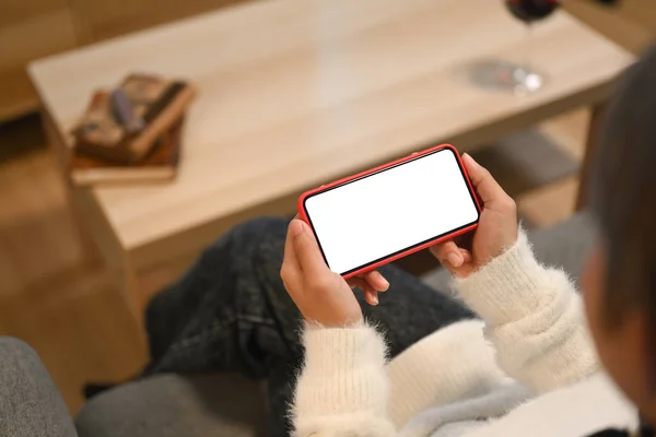 Cropped shot of woman watching media in a smart phone and sitting on a couch at home.