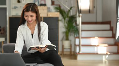 Genç bir kadın elinde bir kitapla kanepede oturuyor ve evinde dizüstü bilgisayarından bilgi okuyor..