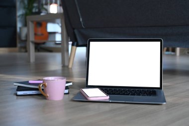 Oturma odasındaki ahşap zemine dizüstü bilgisayar, cep telefonu, kahve fincanı ve defter..