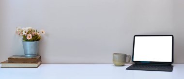 Boş ekran bilgisayar tableti, not defteri, kahve fincanı ve beyaz masadaki vazo ile basit çalışma alanı.