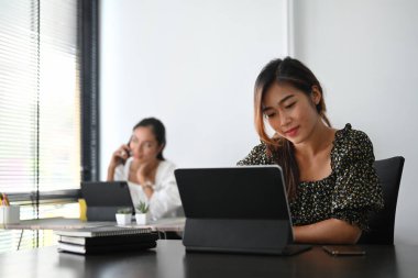 Genç bir kadın bilgisayar tableti üzerinde çalışıyor ve arka plandaki kadın ofiste telefonla konuşuyor..