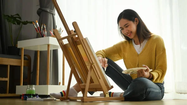 黄色のセーターにパレットを手にした若い女性アーティストが床にキャンバスに絵を描いています — ストック写真