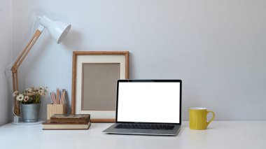 Boş ekran dizüstü bilgisayarı, not defterleri, kahve fincanı ve beyaz masadaki ağaç demliği olan rahat bir çalışma yeri..