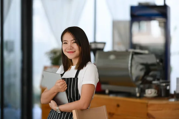 Successful female entrepreneur wearing black apron hands holding tablet and standing in her own coffee shop.
