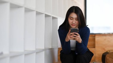 Genç kız öğrenci kütüphanede oturuyor ve akıllı telefon kullanıyor..