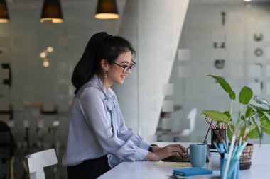Çalışma masasının önünde oturup bilgisayar tabletiyle çalışan neşeli genç bir kadının yan görüntüsü..