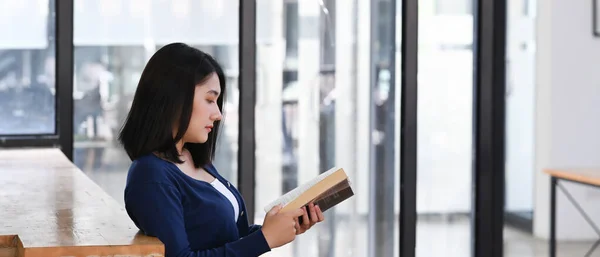 アジアの学生女性の側面図は 現代の図書館に座っている間に本を読んでいます — ストック写真