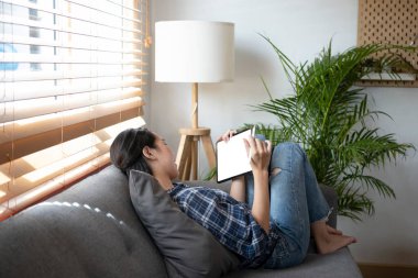 Genç bir kadın rahat koltukta dinleniyor ve evde dijital tablet kullanıyor..