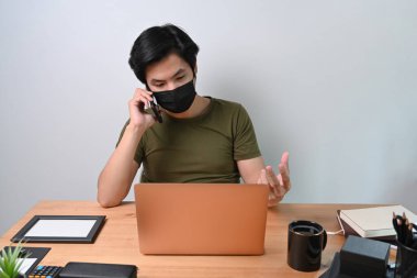 Yüz maskeli genç adam cep telefonuyla konuşuyor ve laptopla çalışıyor..