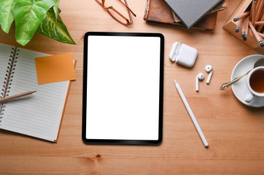 Modern çalışma alanının dijital tablet, kablosuz kulaklık, kahve fincanı ve ahşap masadaki defter ile genel görüntüsü..