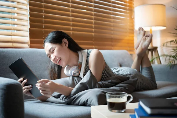 Cheerful young woman lying on couch and using digital tablet.
