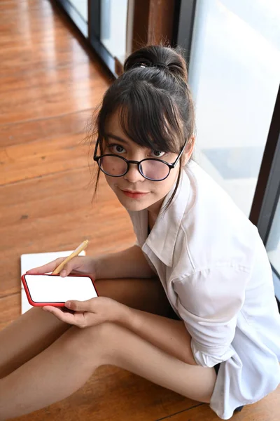身着休闲装 手持智能手机 坐在木制地板上看着相机的年轻女子的画像 — 图库照片