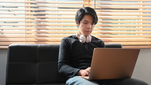 Asian man sitting on couch and surfing internet with laptop computer.