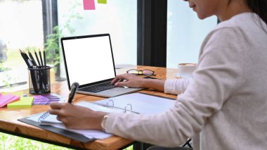 Dizüstü bilgisayarla çalışan ve deftere not alan kırpılmış kadın tasarımcı..