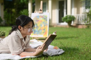 Genç bir kadın çimlerde bir battaniyeye uzanmış, güneş parlıyor ve kitap okuyor..
