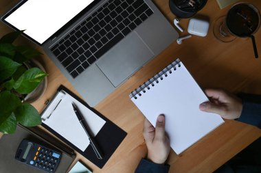 İş adamı elinde defter tutuyor ve tahta masada dizüstü bilgisayar kullanıyor..
