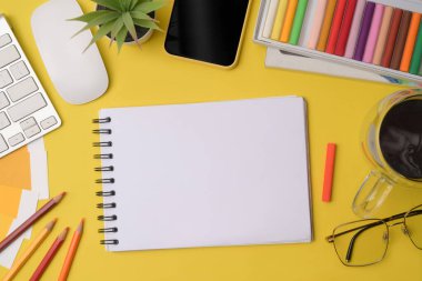 Boş defter, cep telefonu kahve fincanı ve sarı arka planda renkli boya kalemleri..