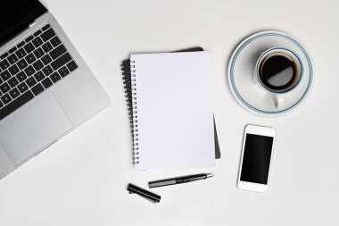 Boş defter, cep telefonu, kahve fincanı ve dizüstü bilgisayar beyaz masada..