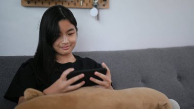 Oturma odasında koltukta otururken cep telefonu kullanan gülümseyen Asyalı kız..