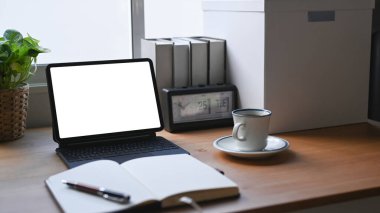 Bilgisayar tableti, defter, kahve fincanı ve tahta masadaki kitaplarla rahat çalışma alanı.