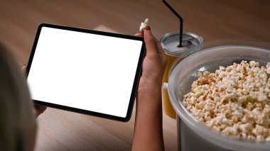 Genç bir kadın dijital tablette online film izliyor ve evdeki kanepede patlamış mısır yiyor..