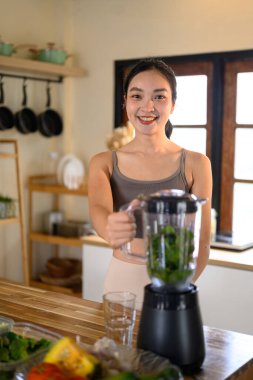 Gülümseyen kadın mutfaktaki mikserde sağlıklı yeşil bir meyve püresi hazırlıyor..